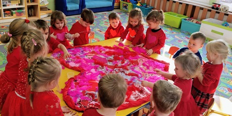 Powiększ grafikę: Walentynki - dzieci w czerwonych ubraniach wyklejają duże papierowe serce ozdobami: piórami, cekinami brokatem, naklejkami